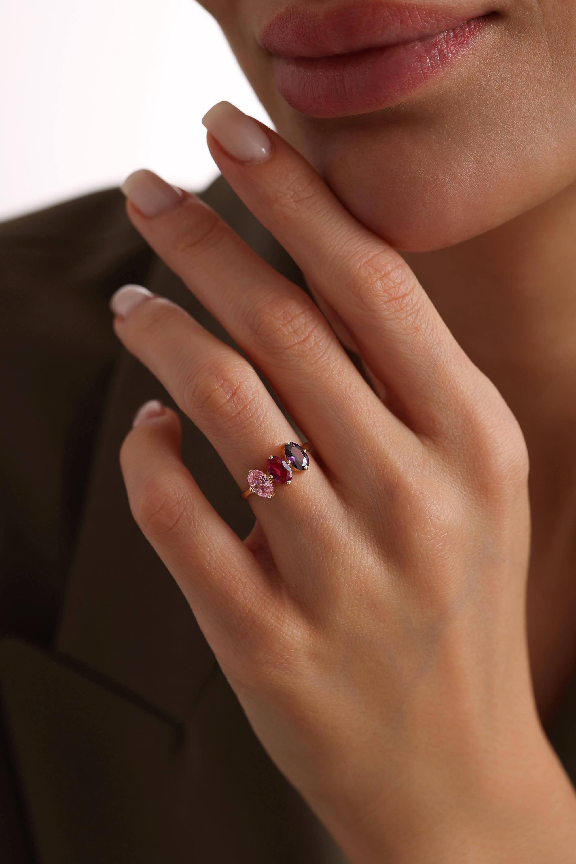 May include: A gold ring with three oval-shaped gemstones. The gemstones are pink, red, and purple.