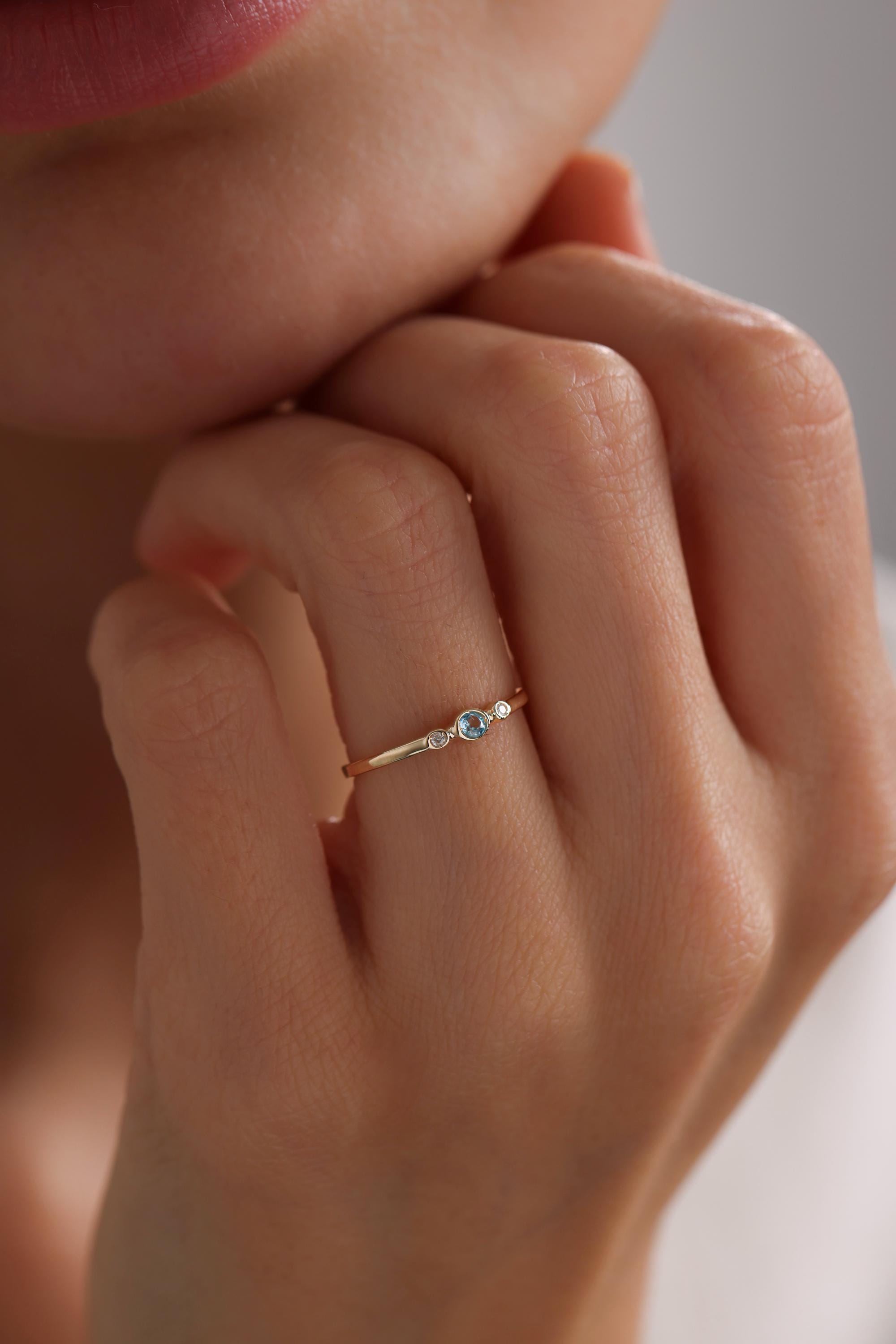 May include: A delicate gold ring with three small round gemstones. The center stone is a larger, light blue gemstone, and the two stones on either side are smaller and white.