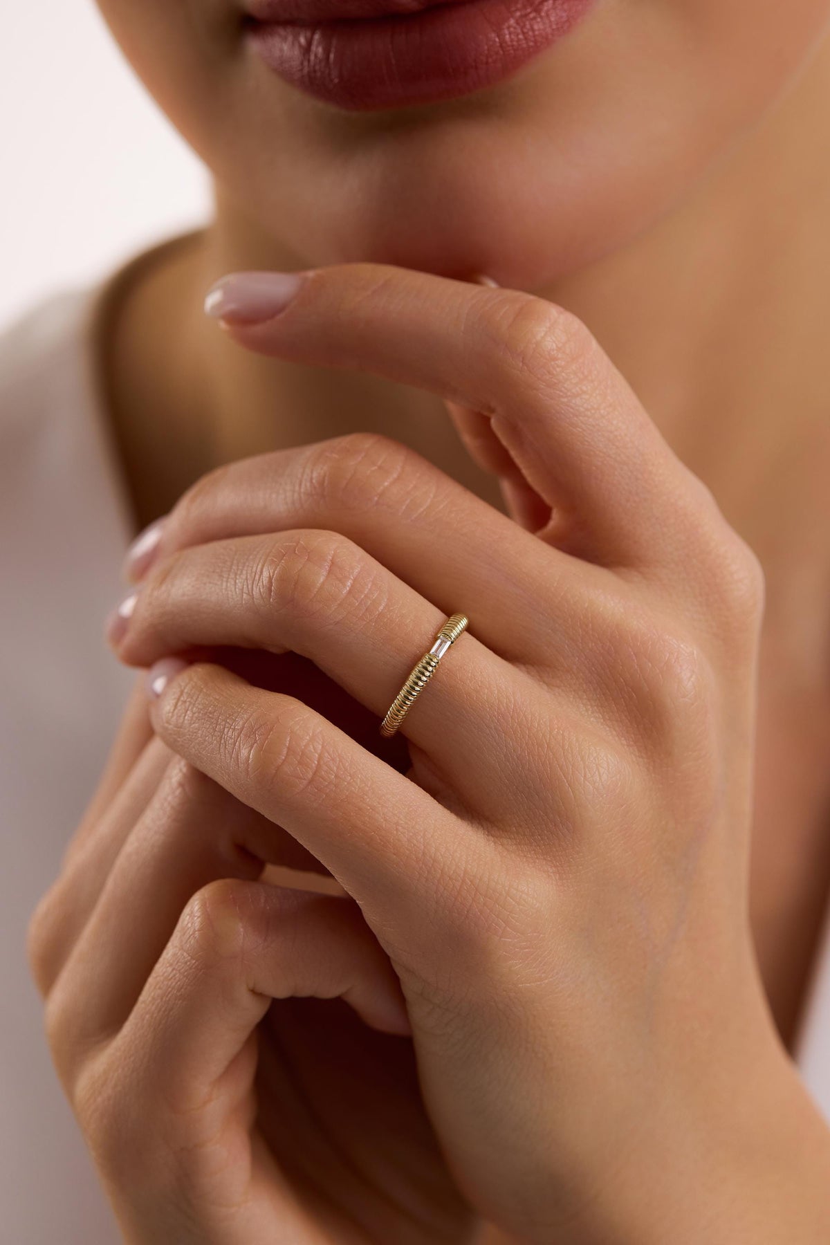 May include: A gold ring with a textured band and a small, rectangular, clear stone. The ring is worn on a finger, showcasing its delicate design and elegant simplicity. The ring's band has a ribbed texture.