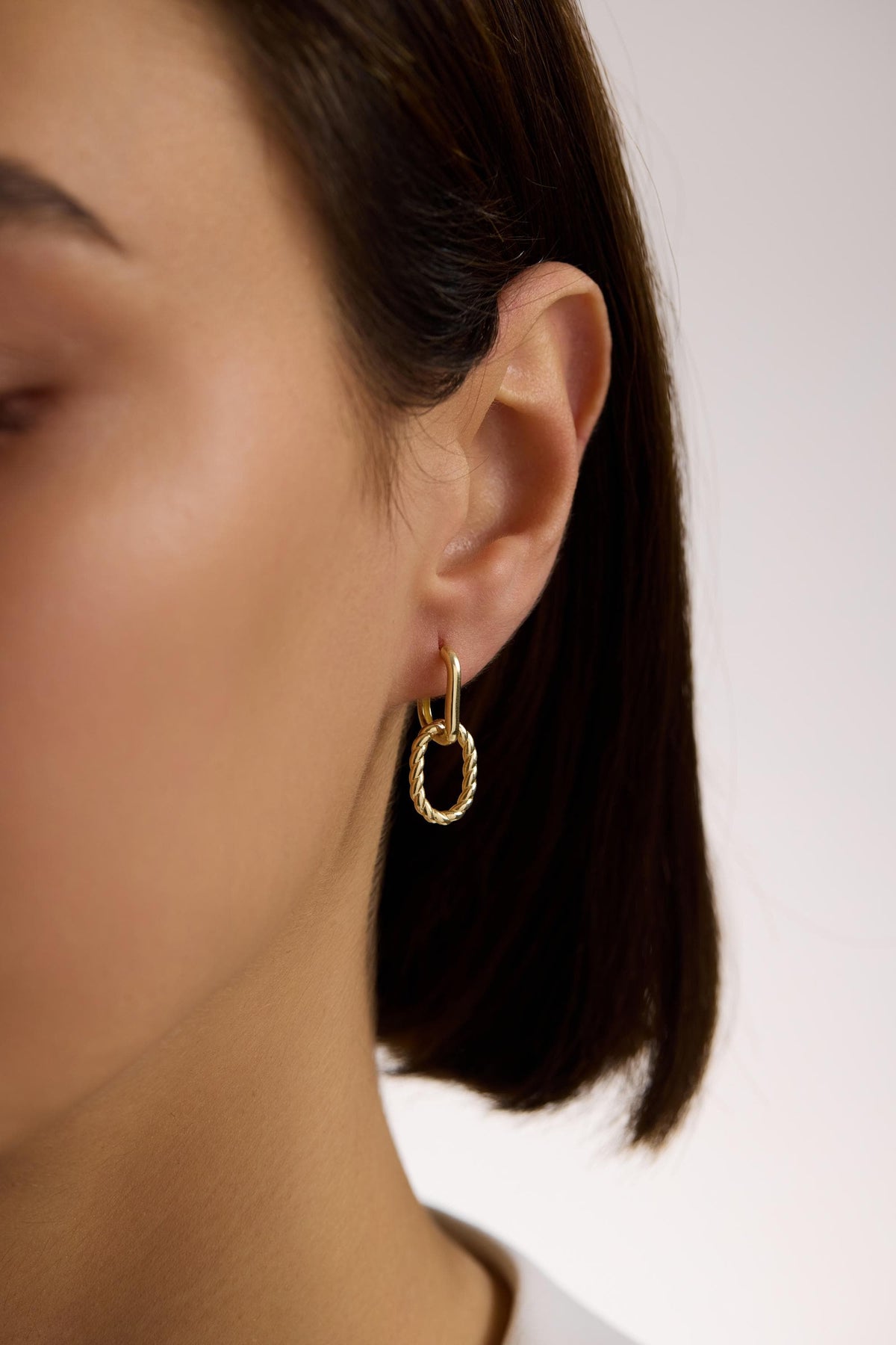 May include: Gold-toned hoop earrings with a textured oval pendant. The earrings are connected by a rectangular link. The oval pendant has a twisted rope design. The earrings are worn on the ear.