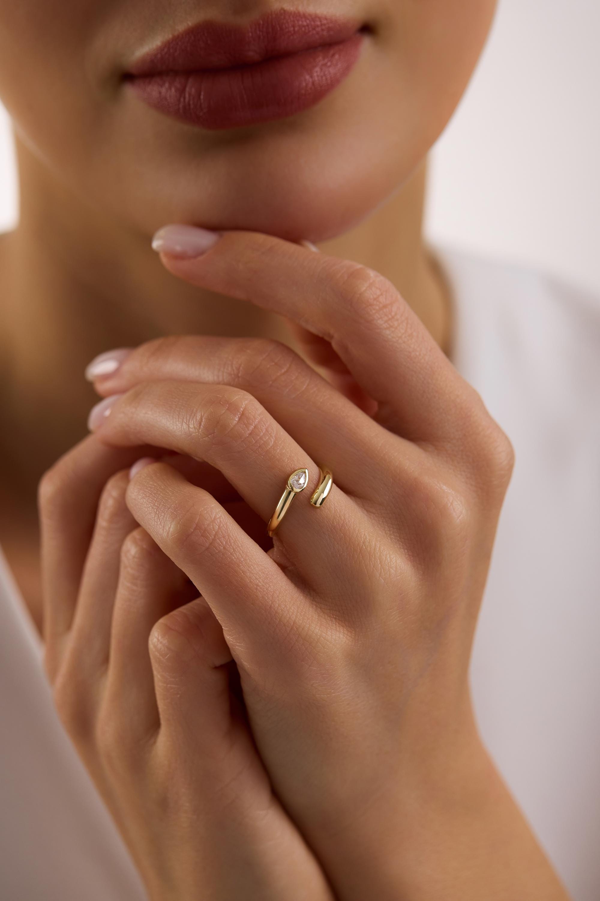 May include: A gold-colored ring with a teardrop-shaped clear gemstone. The ring has an open design, with the band splitting to cradle the gemstone. The ring is worn on a finger.